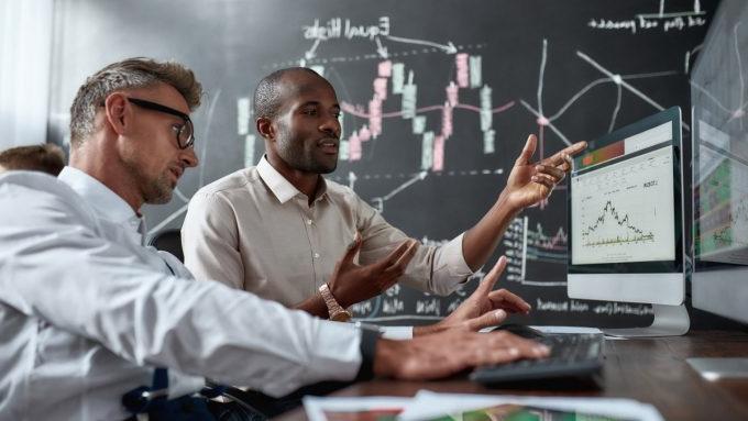 Two diverse colleagues traders talking to each other, looking at graphs while sitting in the office in front of multiple computer screens. Stock trading, people, business concept.